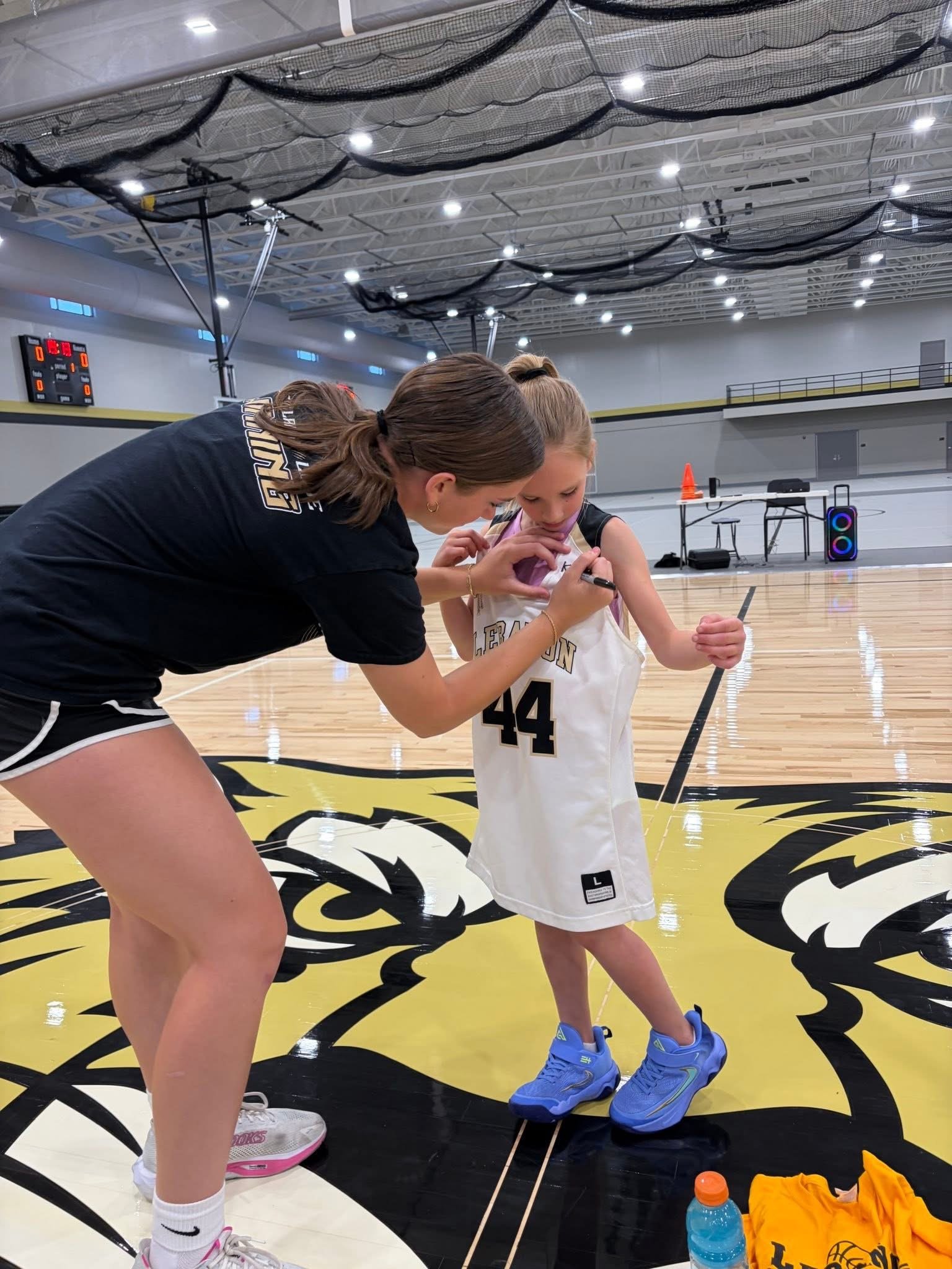 A young girl wearing a basketball uniform with the number 44 on it is standing on a basketball court while a woman, possibly her coach, assists her with a practice drill. The woman is leaning over, guiding the girl who is holding a pen and marking something on her arm. The gym has a yellow and black lion logo on the floor, and in the background, there are tables, chairs, a speaker, and a water bottle.