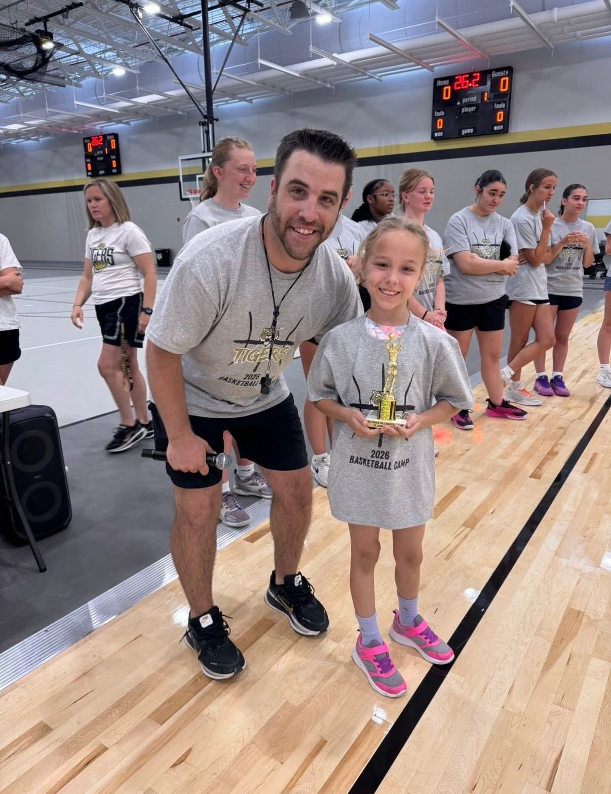 A young girl holding a trophy at a basketball camp, smiling, standing next to an adult man in a gymnasium with a group of girls in the background.