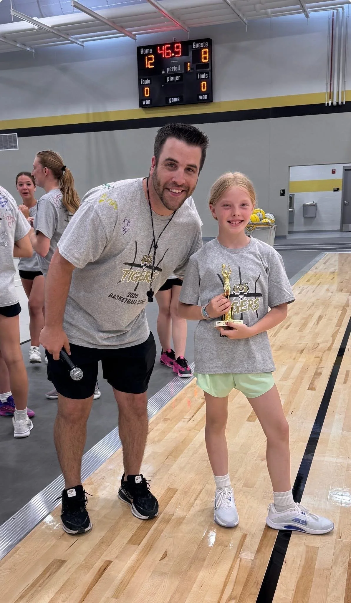 A young girl in sportswear holding a small trophy, standing next to a man with a microphone in a gymnasium. The girl is smiling and the man is smiling at the camera.