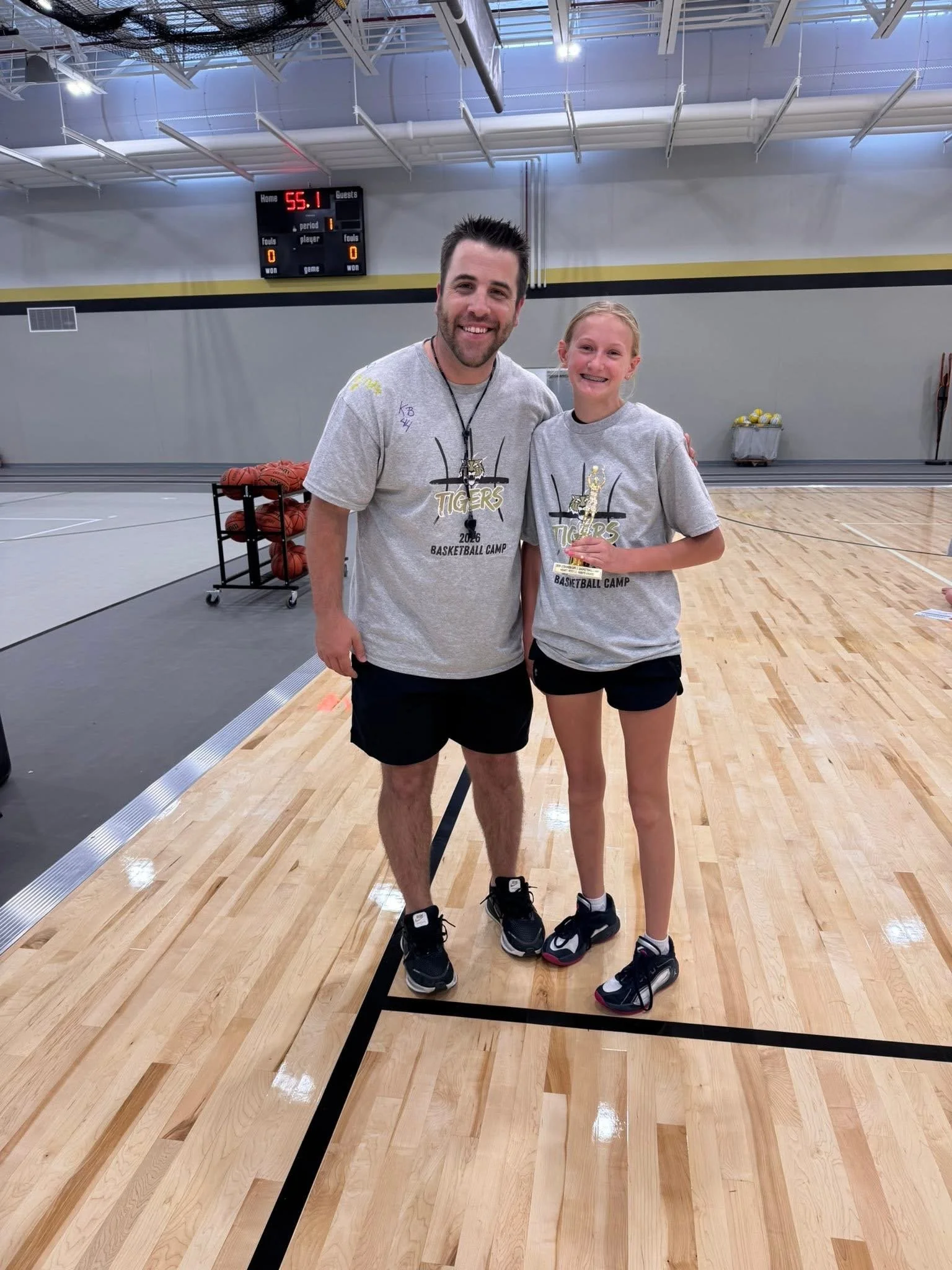 A man and a girl standing on a basketball court inside a gym, smiling and posing for a photo, wearing gray T-shirts and athletic shoes.