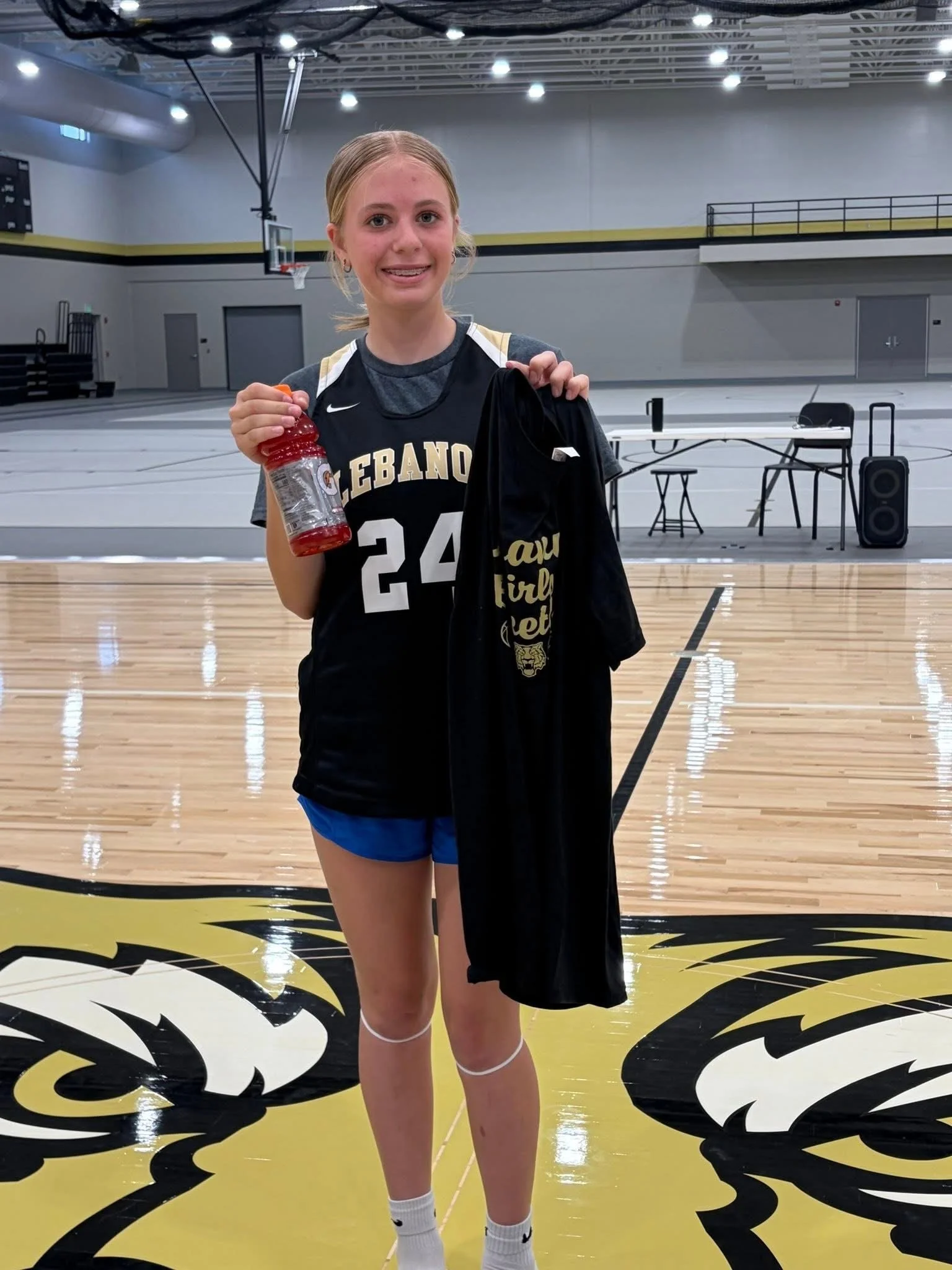 A young girl is standing in an indoor basketball court, holding a water bottle and a jacket. She is wearing a black basketball jersey with the number 24 and the word 'Lebanon' on it, along with blue shorts and white Nike socks. The background shows basketball hoops and a stage area.