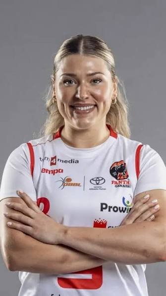 A woman in a white sports jersey with various logos, smiling with her arms crossed.