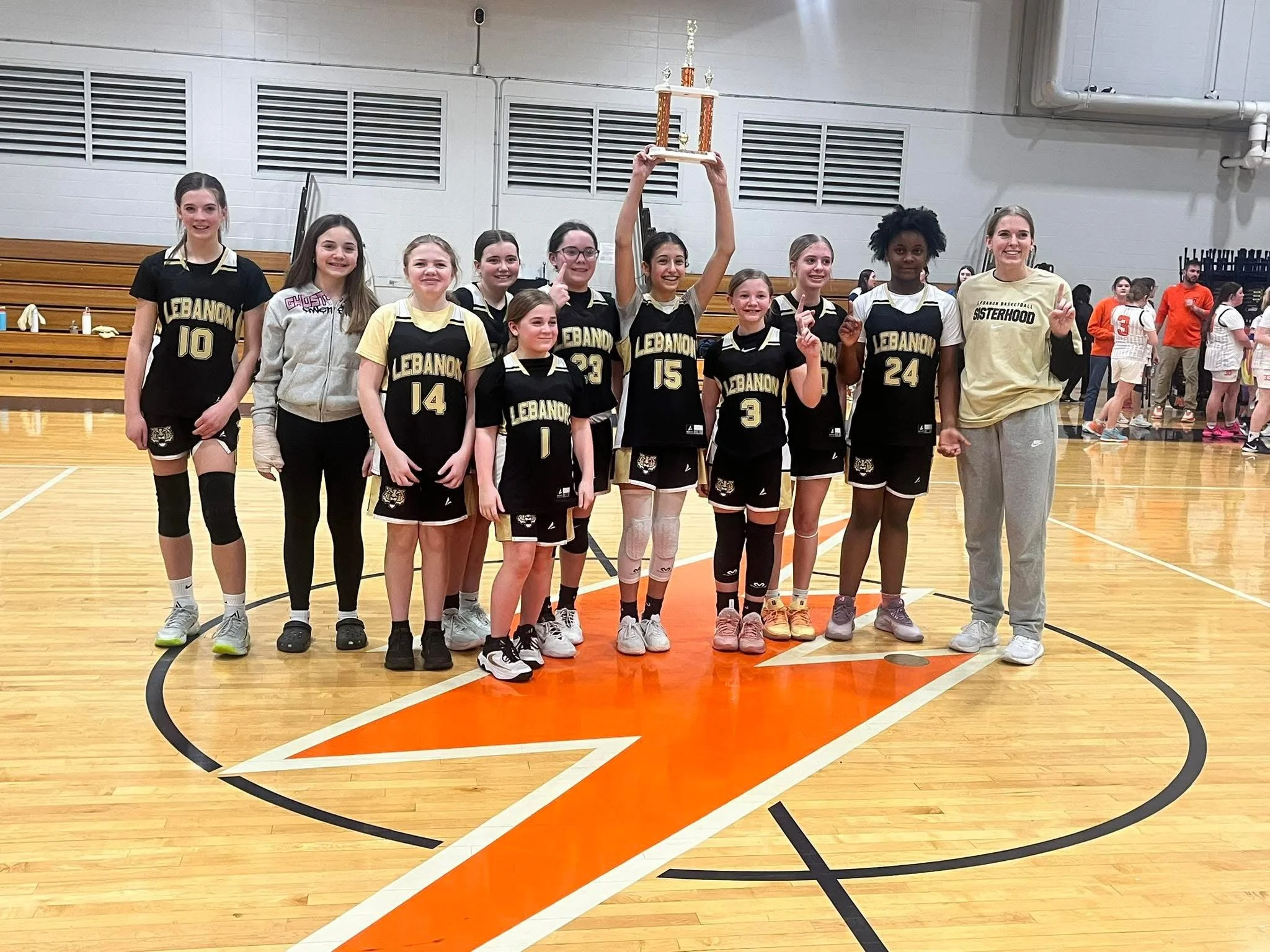 A girls' basketball team celebrating with a trophy in a gymnasium after a game or tournament, with their coach standing to the right.