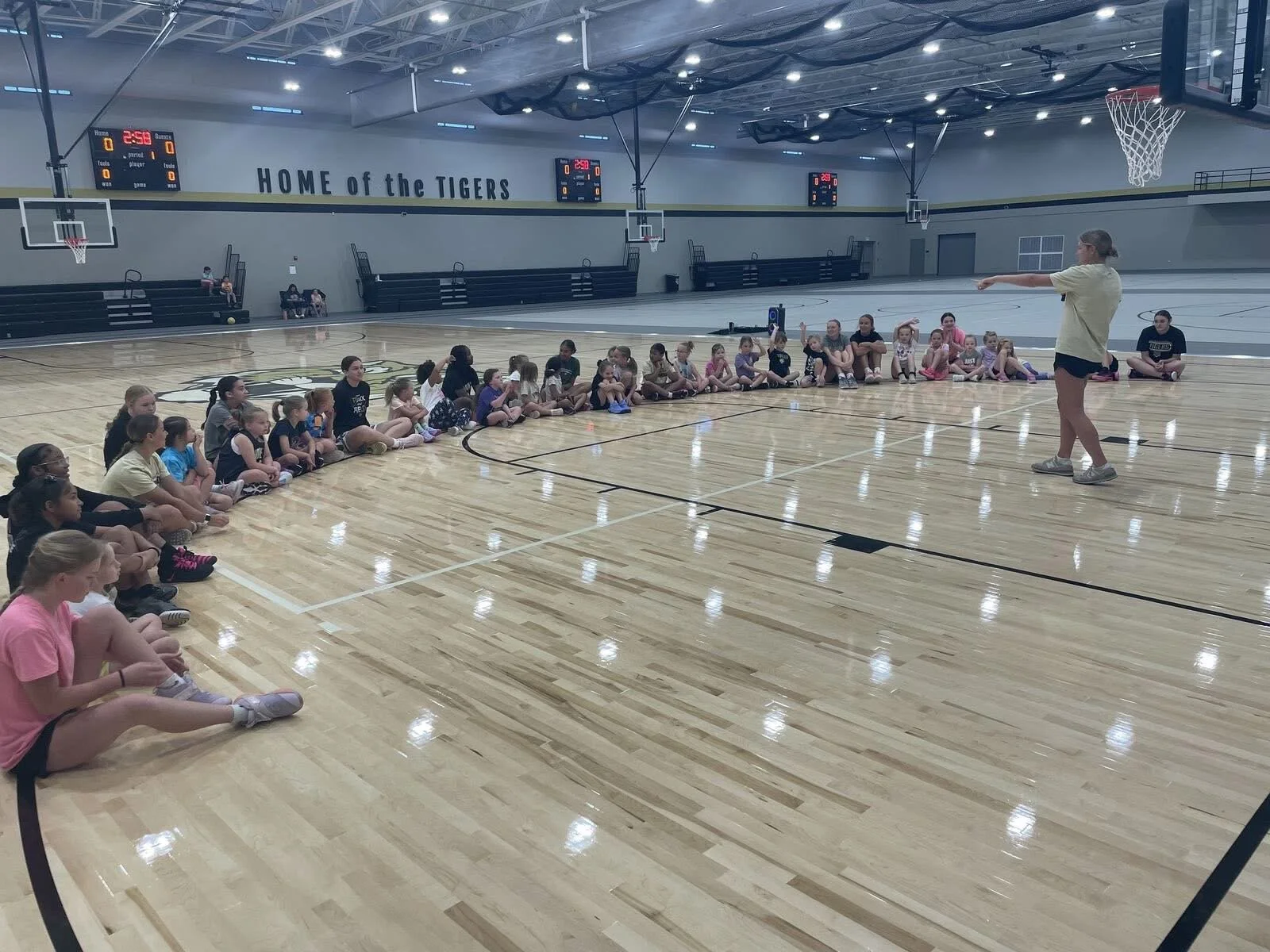 A woman is giving instructions to a group of children seated on the shiny wooden floor of a basketball gym. The gym has basketball hoops, scoreboards, and the phrase "HOME of the TIGERS" painted on the wall.