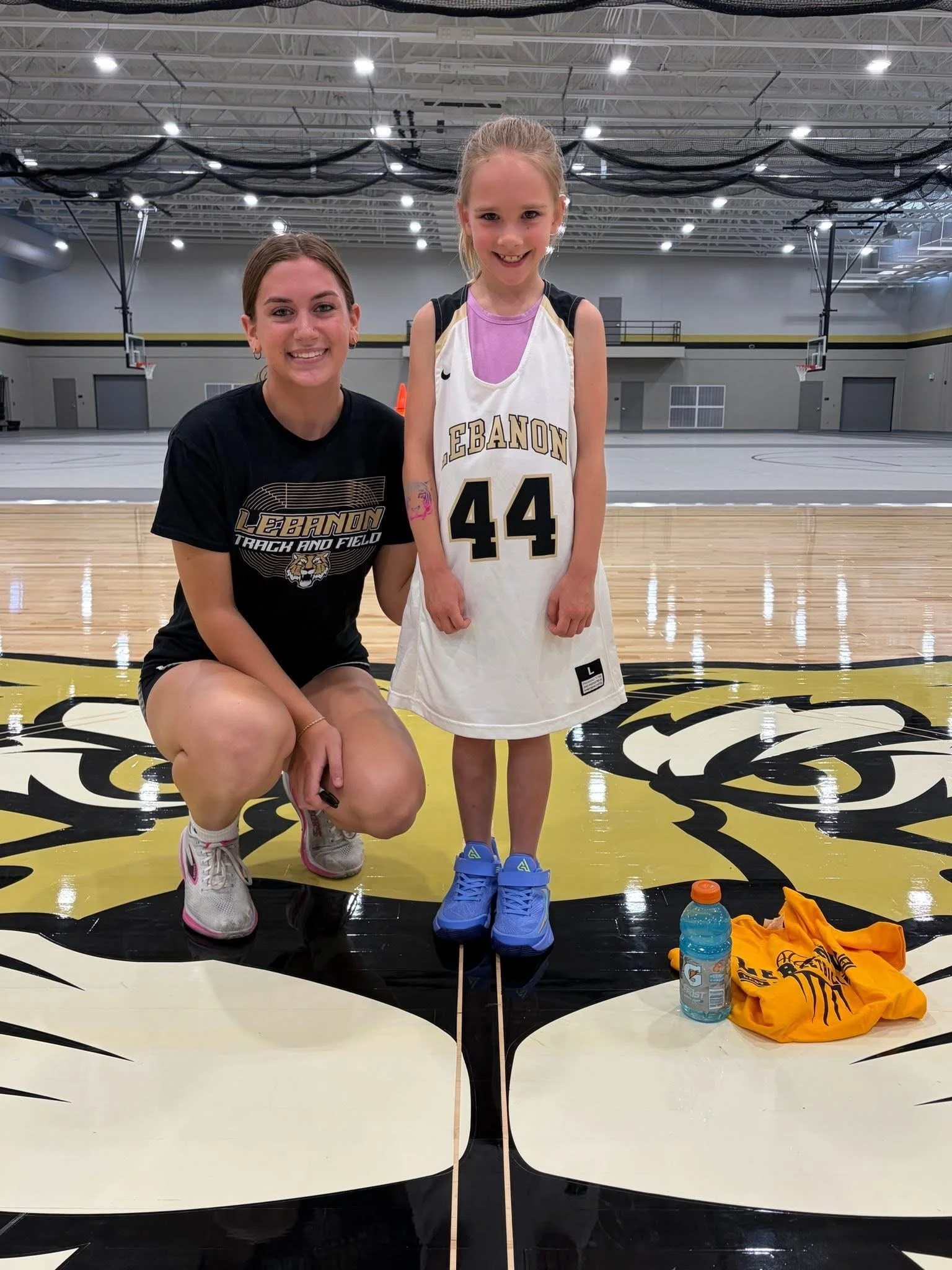 A young girl with a bright smile wearing a Lebanon basketball jersey is standing inside a gymnasium with an older girl kneeling next to her, both smiling at the camera. The gym has a polished wooden court, basketball hoops, and black netting hanging from the ceiling. In the foreground, there are water bottles and an orange shirt on the floor.