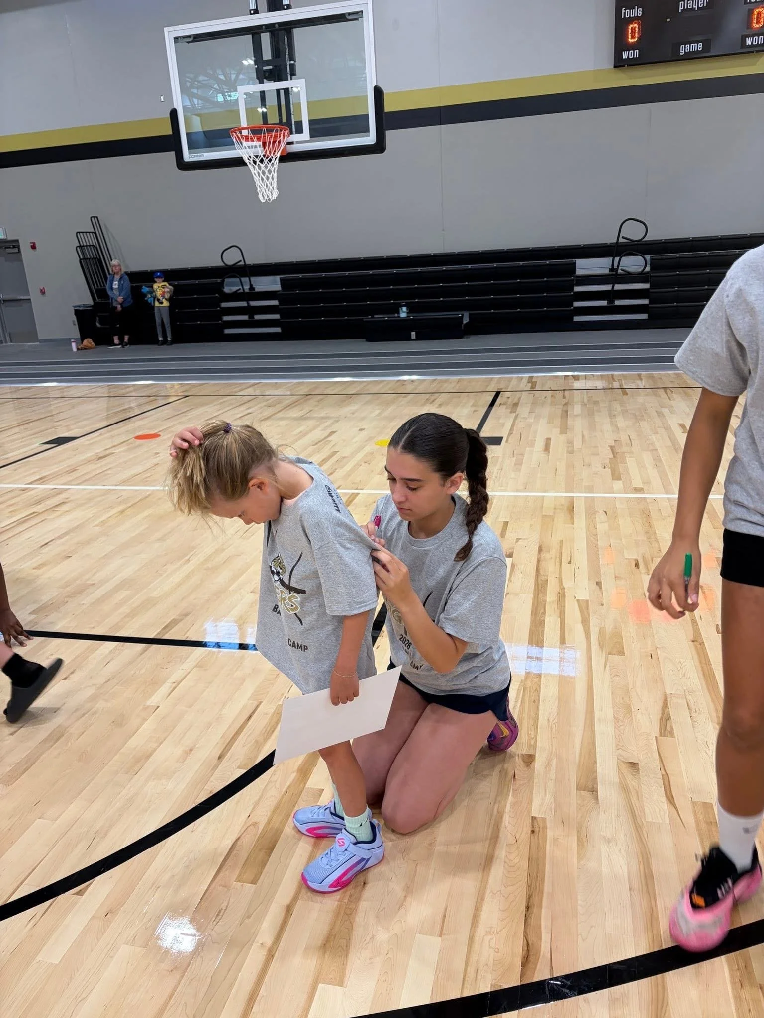 Two girls wearing gray t-shirts and shorts, one kneeling and the other standing, in a gymnasium with wooden flooring, while other children are partially visible around them.