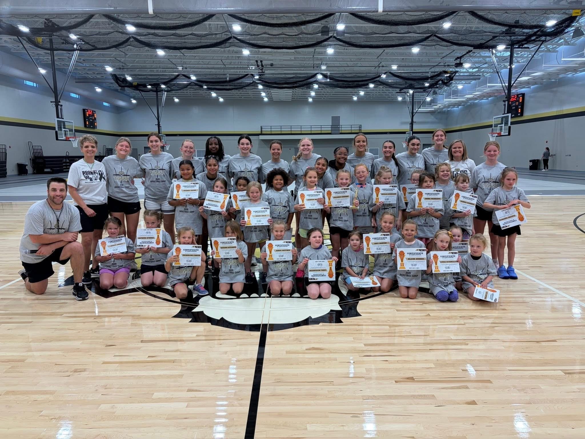 A group of children and adults in a basketball gymnasium holding certificates, posing for a photo. The gym features a basketball court, scoreboard, and black nets hanging from the ceiling.