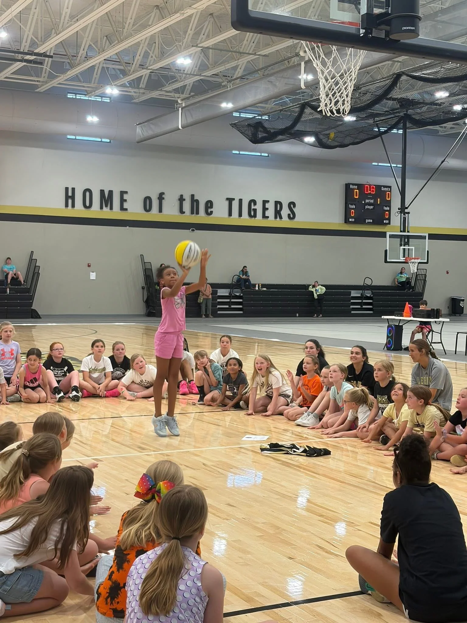 A girl in a pink outfit is jumping to shoot a volleyball in a gymnasium during a youth sports activity. She is surrounded by children seated on the floor, watching her. The gym has a basketball hoop, scoreboard, and a wall with the words 'HOME of the TIGERS' displayed.
