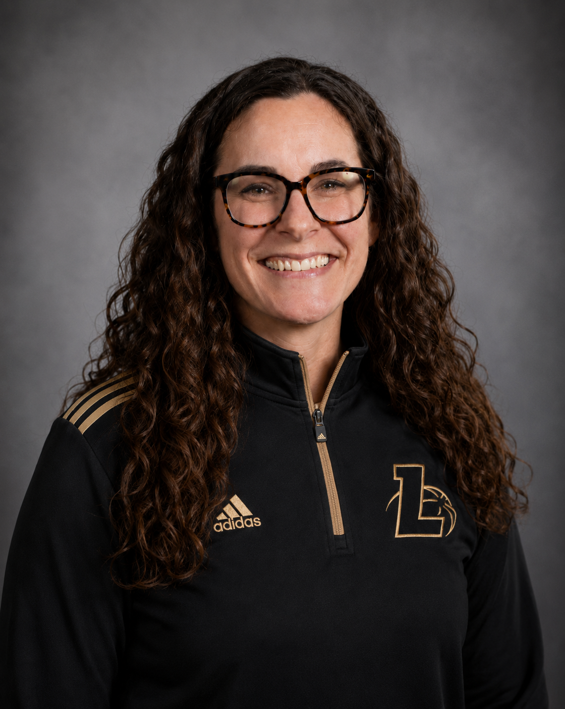 A woman with long curly hair, wearing glasses and a black sports jacket with gold accents, smiling at the camera against a gray background.
