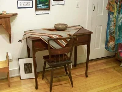 A small wooden desk with a striped cloth runner, a ceramic bowl on top, and a wooden chair in front. Wall decor includes framed artwork and a shelf with plants, with a door and colorful curtains nearby.