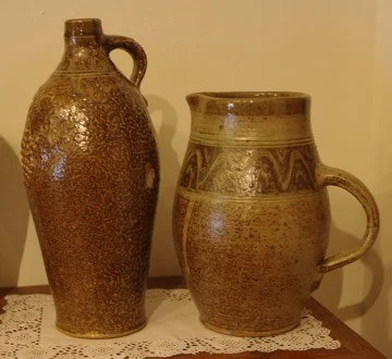 Two brown ceramic vessels, one tall and narrow with a handle, the other shorter with a handle and decorative bands, placed on a lace-covered surface.