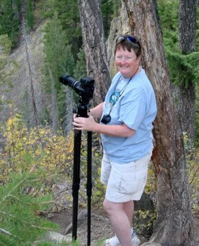 Woman with short hair and glasses holding a telescope outdoors in a forested area with trees and greenery. Meg Gouraud