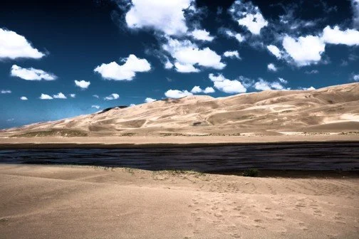 Desert landscape with sand dunes, a dark river or stream, and a partly cloudy blue sky.