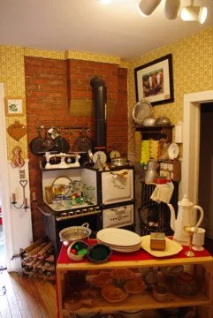 Kitchen with vintage appliances, dishes, and cookware on shelves and counters, brick wall, yellow wallpaper, and framed picture