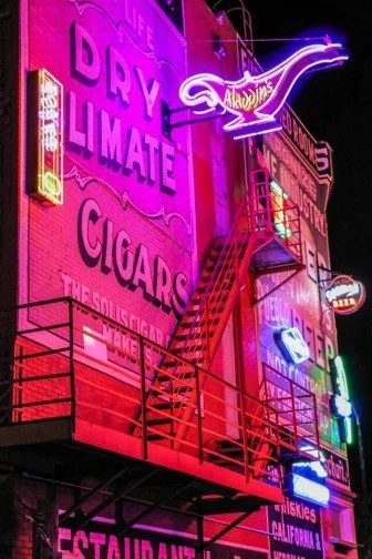 Neon signs on a building advertising cigars, liquor, and a restaurant, illuminated in vibrant pink, purple, and blue colors.