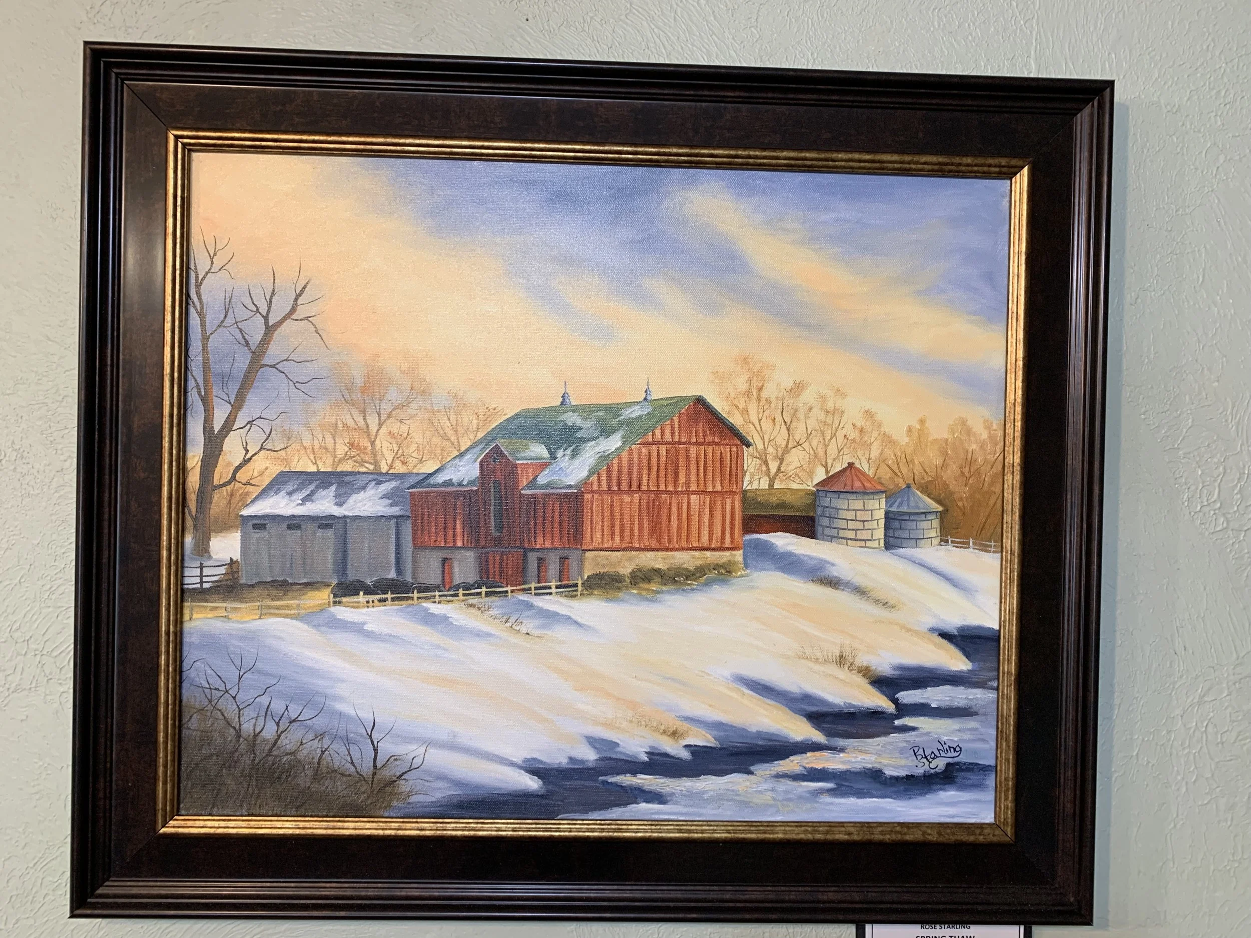 A framed painting depicting a winter farm scene with snow-covered fields, a red barn, silos, leafless trees, and a colorful sky at sunset.