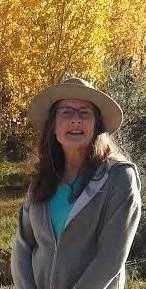 A woman wearing a large sunhat, glasses, and a gray jacket standing outdoors with trees showing autumn foliage in the background. Jan Lata