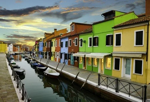 Colorful houses along a canal with boats docked, under a cloudy sky at sunset in Burano.