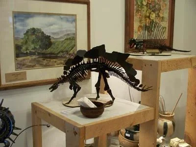 A dinosaur skeleton model displayed on a wooden table with framed landscape paintings and art supplies in the background.
