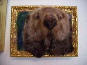 A wool felted sculpture of an otter displayed in a decorative gold frame on a wall.