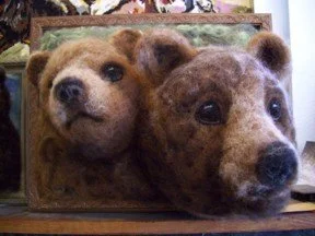 Two realistic bear head sculptures made of wool, one with a puppy-like appearance, in a wooden frame.