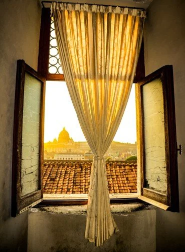 View of a city skyline through an open window with yellow curtains, showing rooftops and a domed building in the distance at sunset.