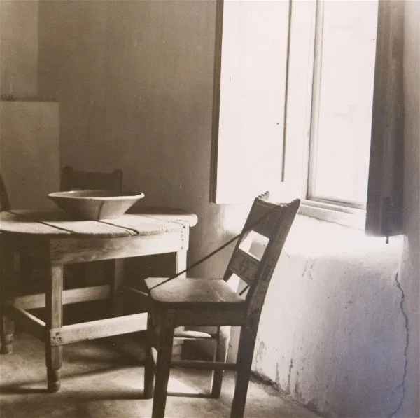 A rustic wooden table with a ceramic bowl, a simple wooden chair, and an open window letting in light, in a cozy, vintage-style kitchen.