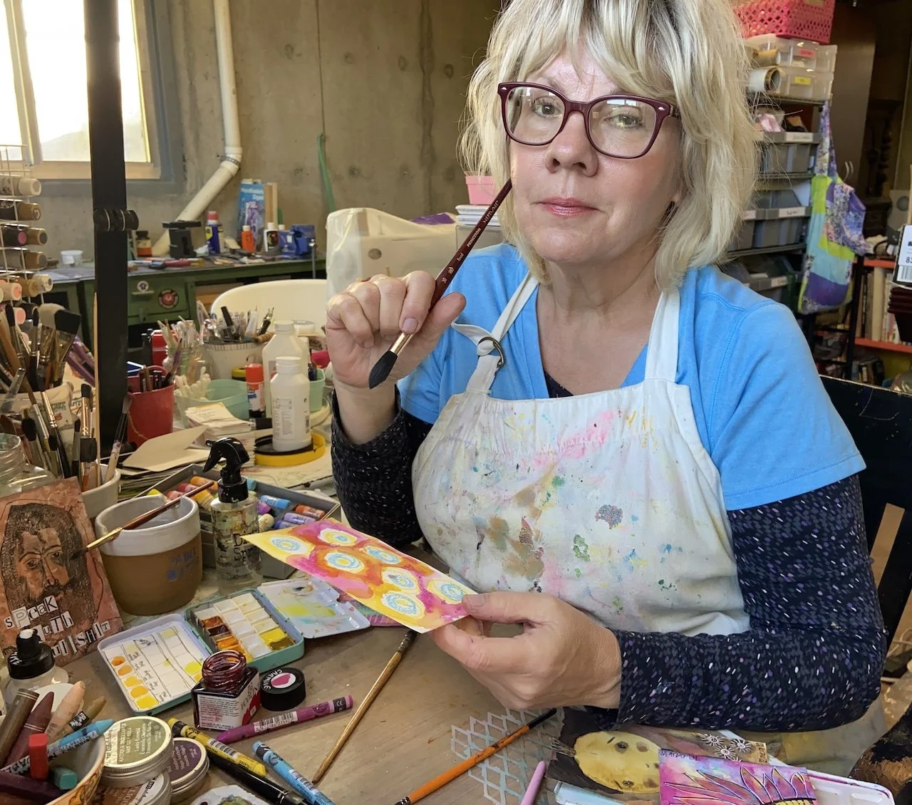 A woman with blonde hair and glasses, wearing a blue shirt and a paint-splattered apron, sitting at a cluttered art table in a studio, holding a colorful painted paper and a paintbrush.