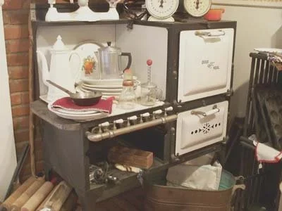 A vintage stove with a white teapot, mixing bowls, and kitchen utensils on top. There are two shelves with kitchen items, and a large rail pipe with hanging utensils beneath the stove. A small oven or storage box is on the right side, and a storage b