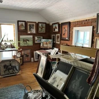 Room filled with framed horse and landscape artwork leaning against walls and furniture, with some art on the wall. There are large windows letting in natural light, and various tables and surfaces covered with art pieces and decorative items.