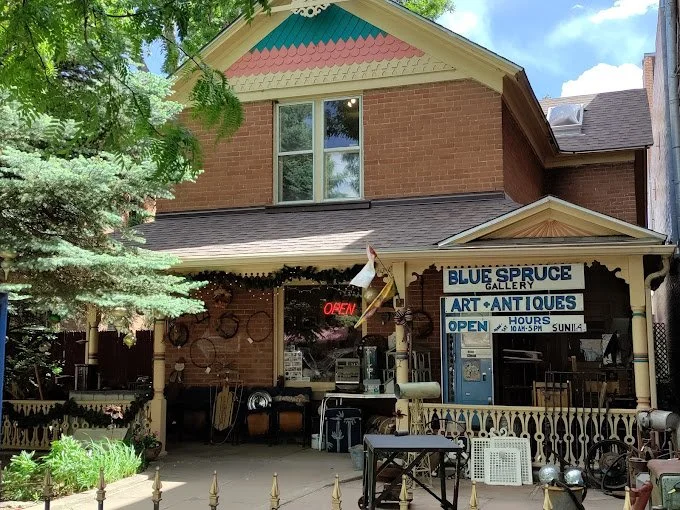 Front of a brick building housing Blue Spruce Gallery, an art and antiques store, with a porch, neon OPEN sign, and decorative roof trim, surrounded by greenery.