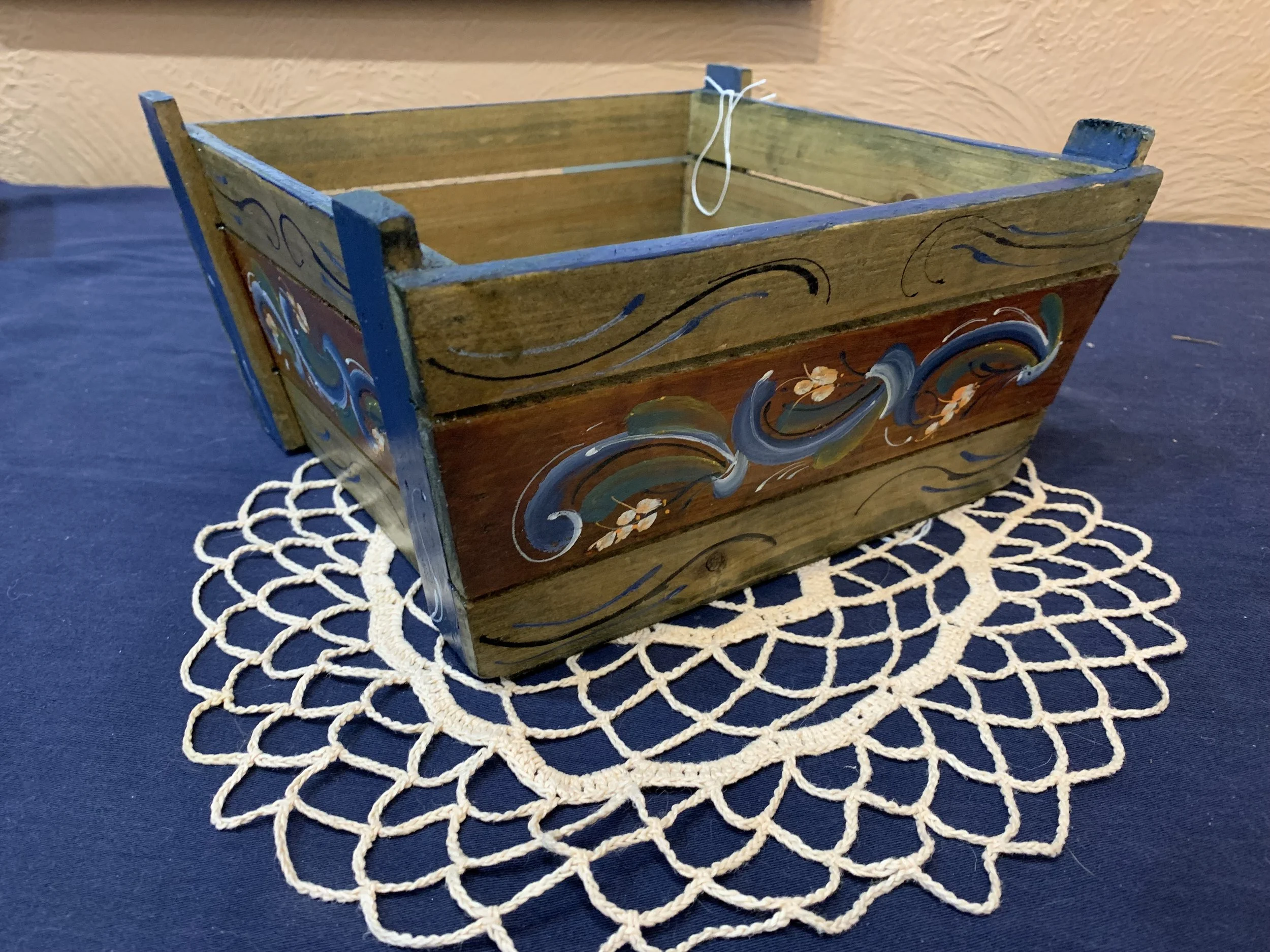 A decorative wooden box with painted designs rests on a white lace doily on a dark blue table. The box has a rectangular shape with open top and painted blue borders and floral patterns.