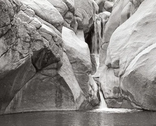 A small waterfall flowing between large smooth rocks into a calm pool of water.