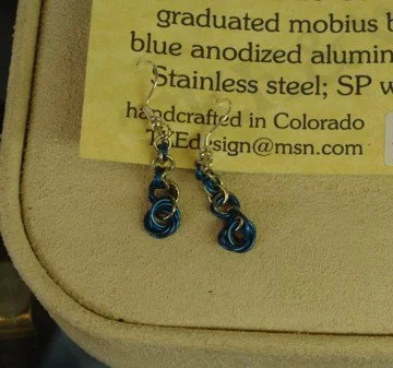 A pair of blue and silver swirl patterned earrings hanging from a hook, on a display stand with a yellow business card in the background.