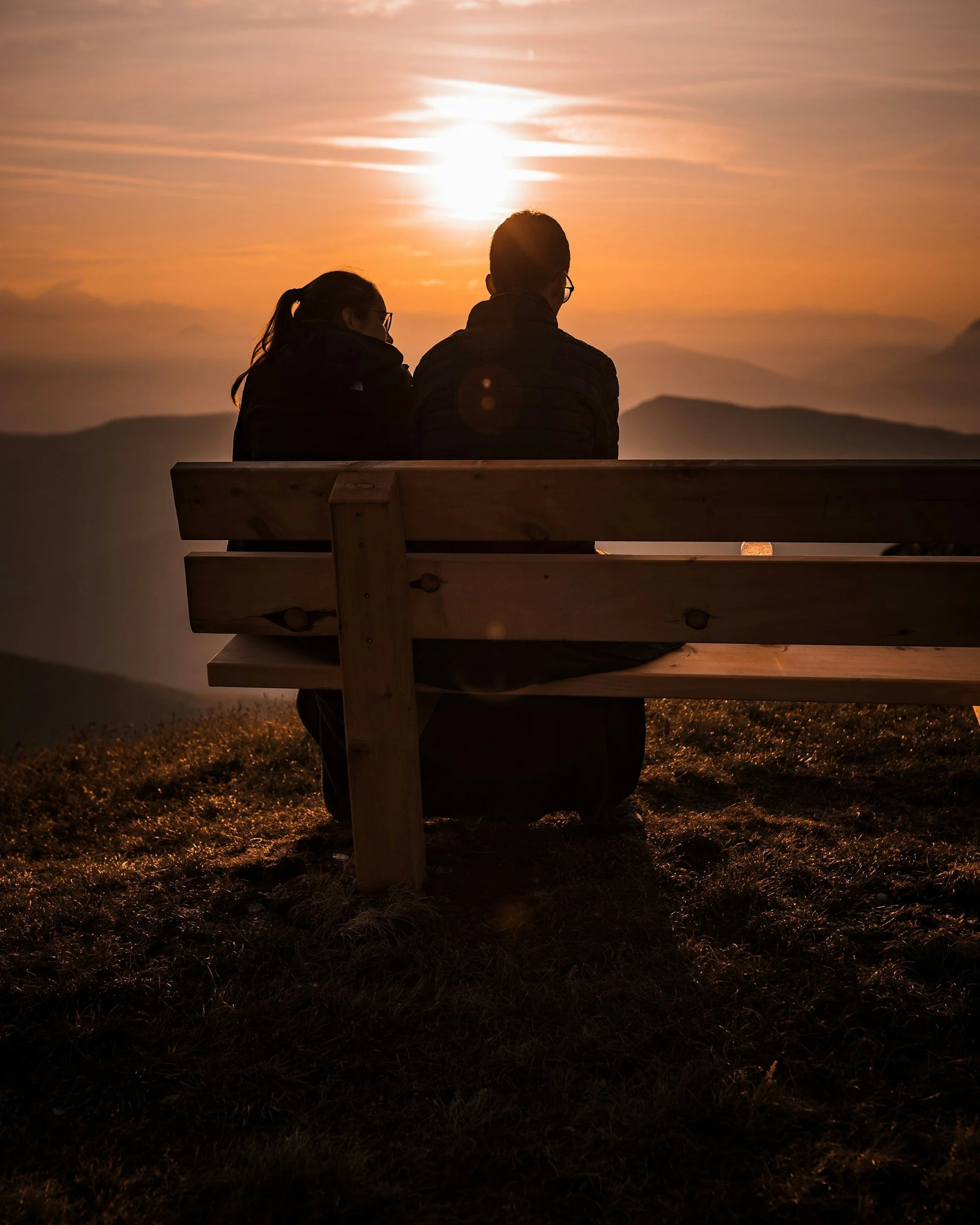 A couple sitting on a wooden bench outdoors during sunset, with mountains in the background and a sky with orange clouds.