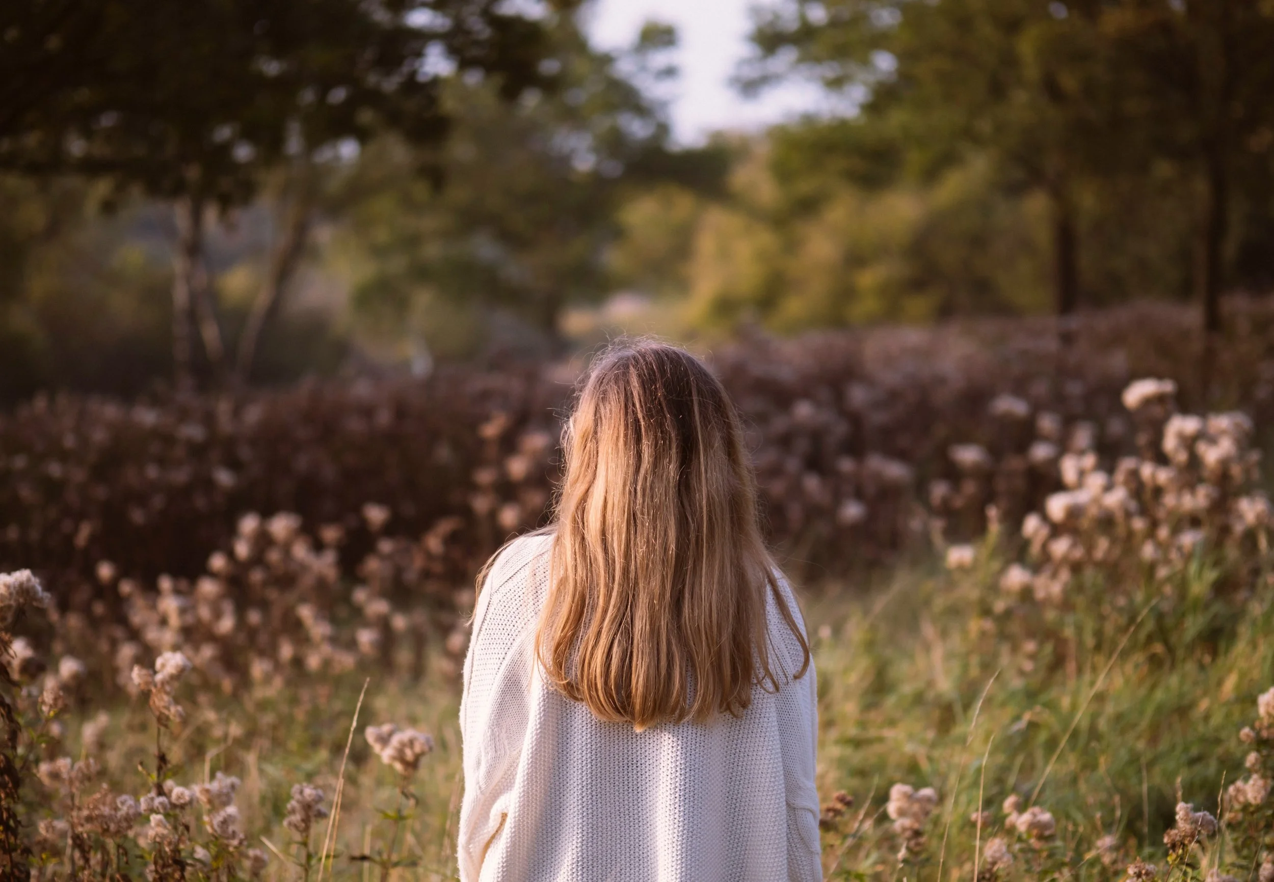 A person with long, light brown hair standing outdoors among tall dry grass and white flowers. The background features blurred trees and a soft, cloudy sky, suggesting a natural, serene setting during late afternoon or early evening.