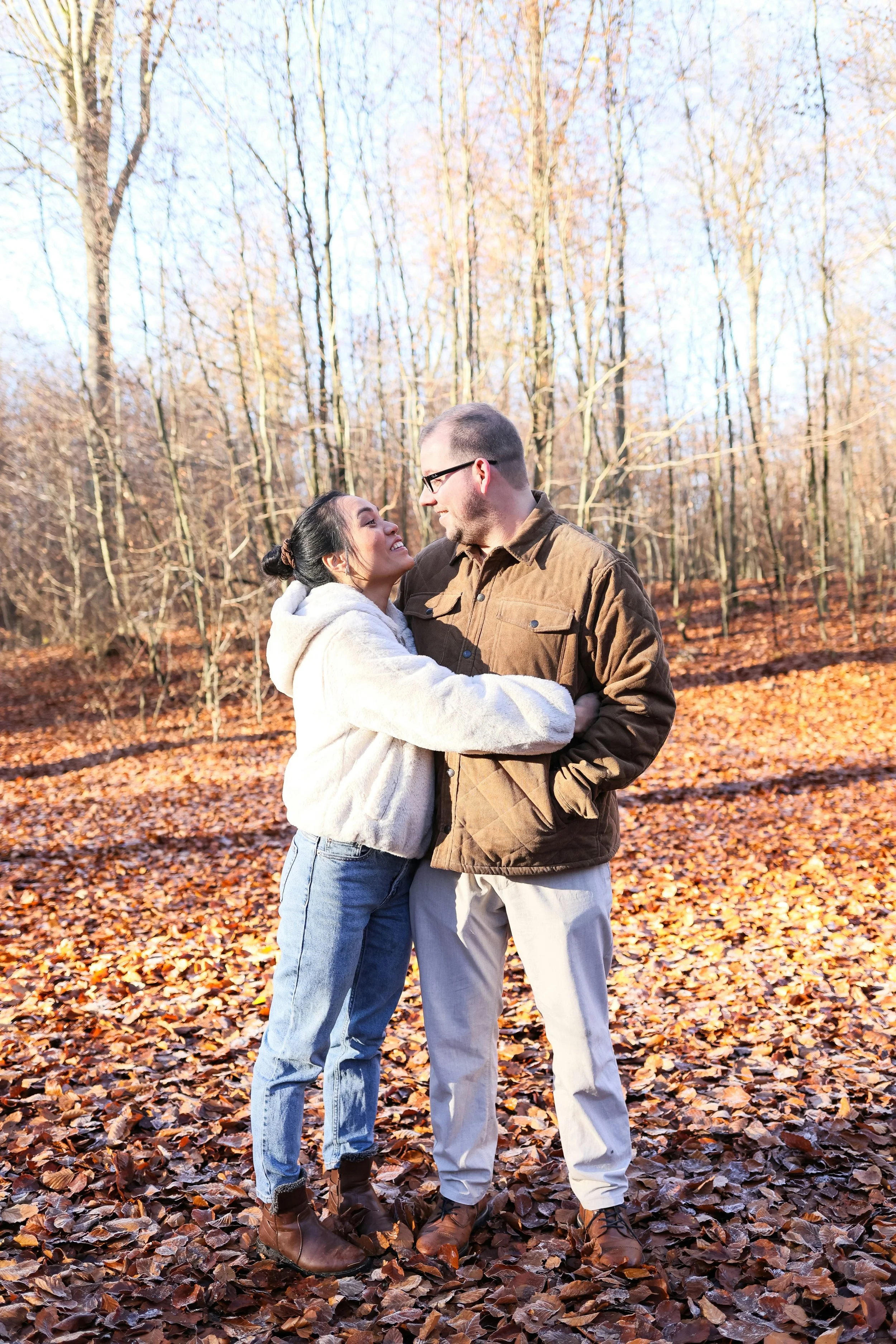 A couple stands close together in a forest with trees and fall leaves on the ground, smiling and looking at each other.
