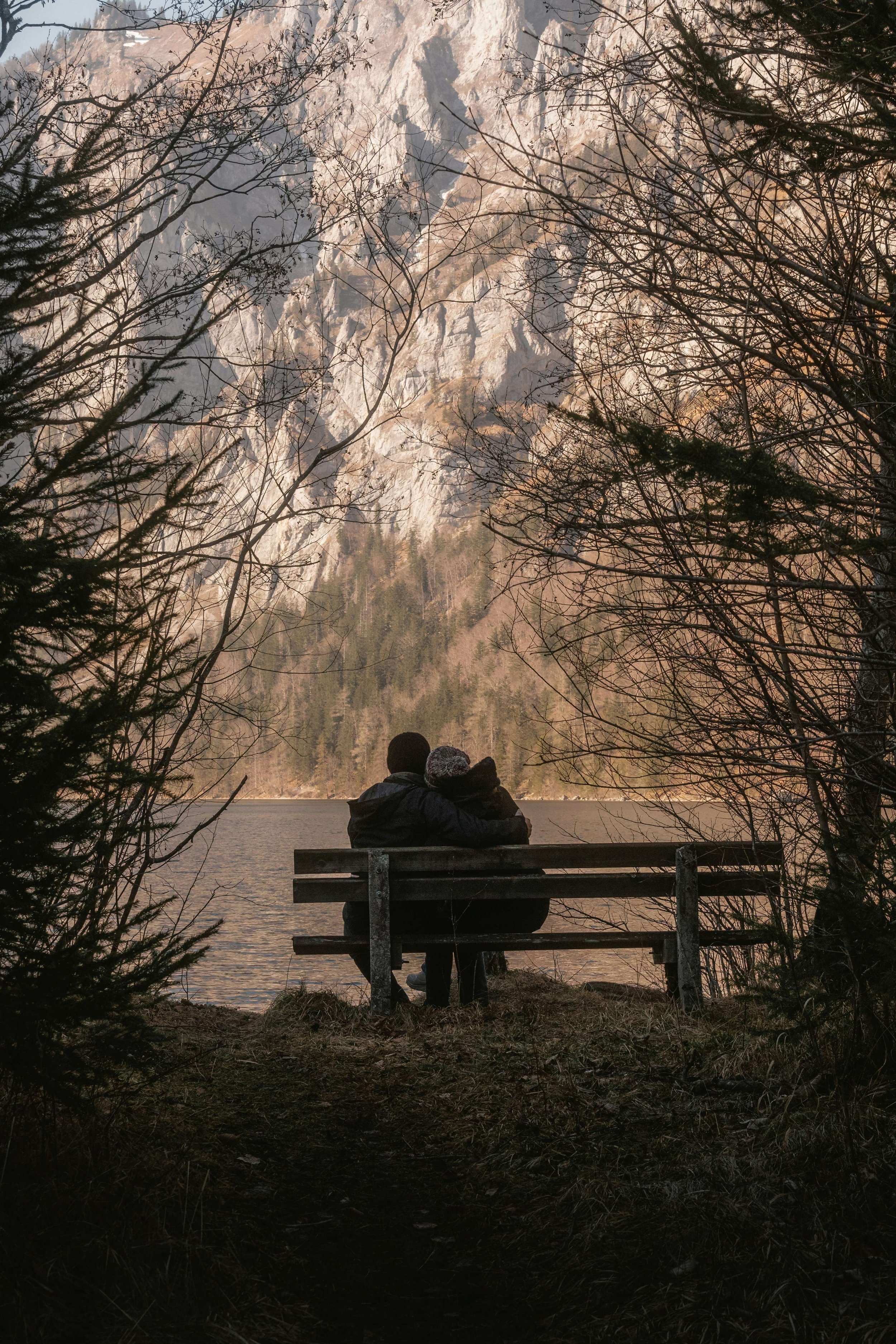 Two people sitting on a park bench by a lake, surrounded by trees with a mountain in the background.