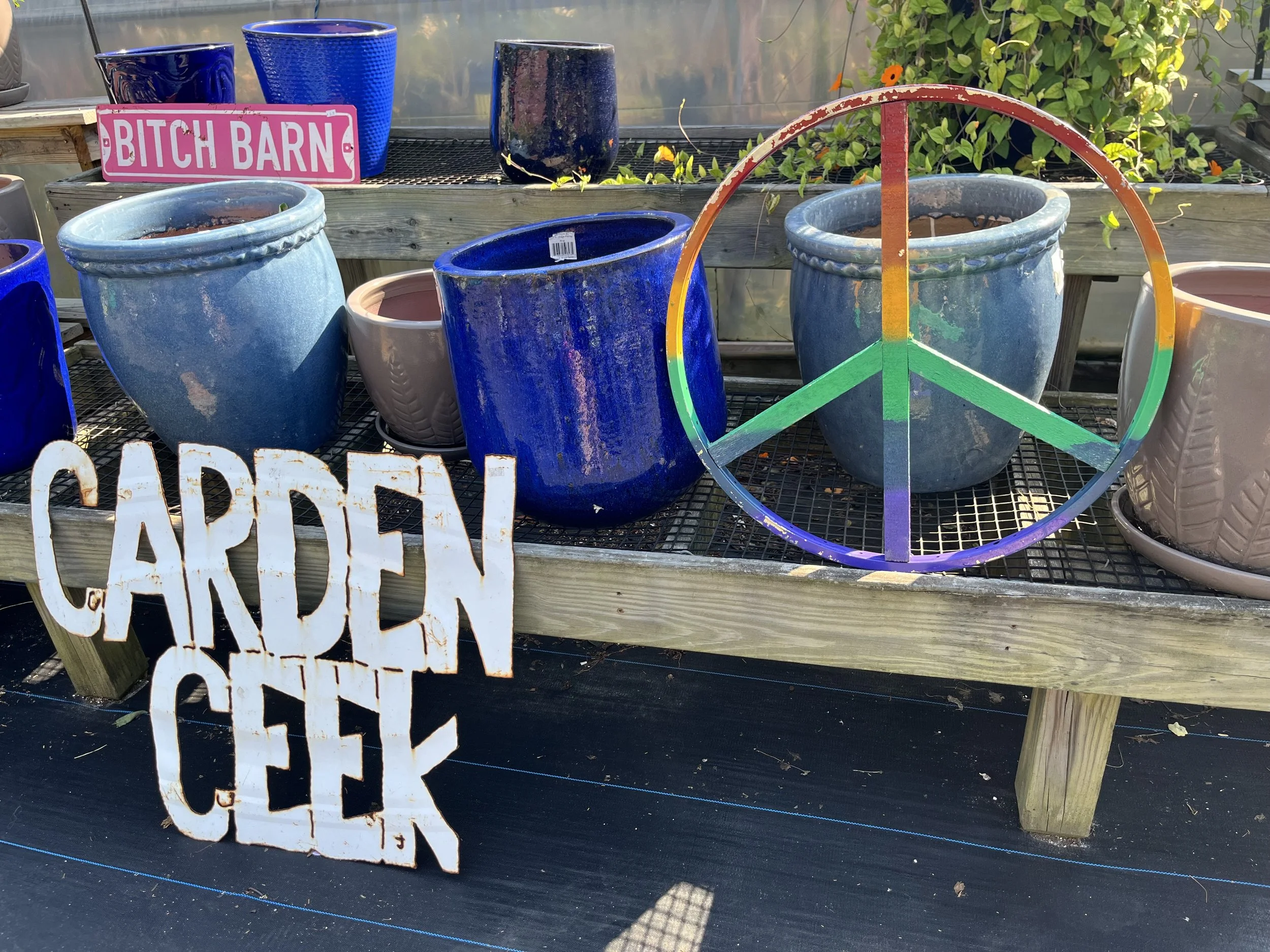 A display of various gardening pots, mainly blue, on a wooden table, with shares of the words 'Garden Center' in white and rust-colored lettering at the bottom left corner. There is also a pink sign in the background that reads 'Bitch Barn'.