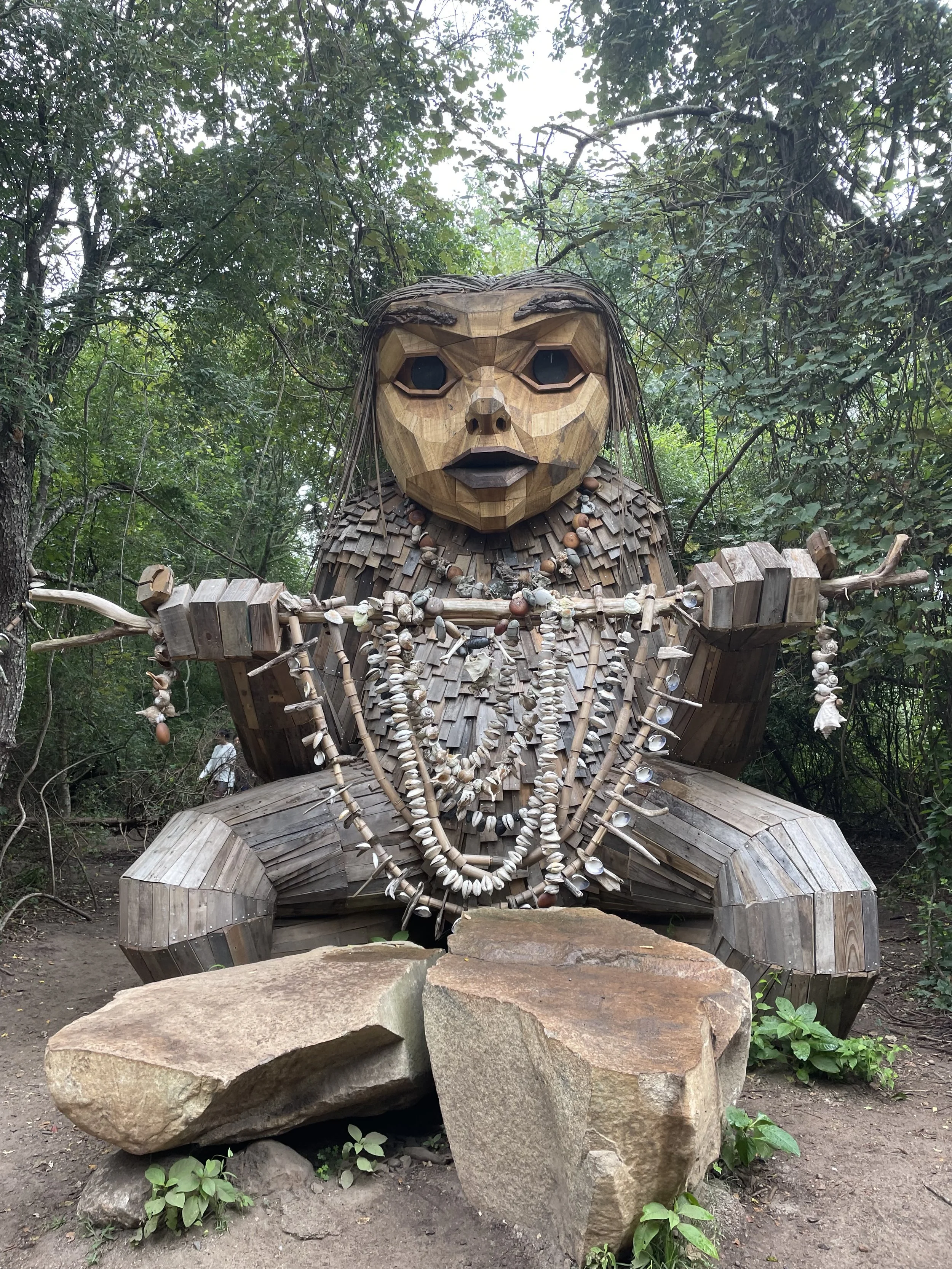 A large wooden sculpture of a woman with detailed facial features, long hair, and necklaces, surrounded by greenery and rocks.