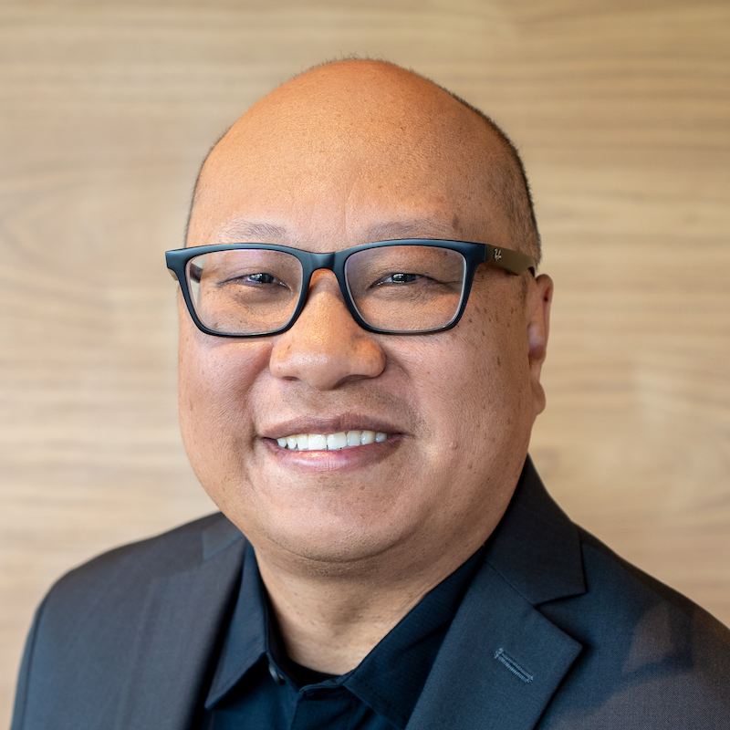 A man with no hair wearing glasses and a black suit, smiling at the camera.