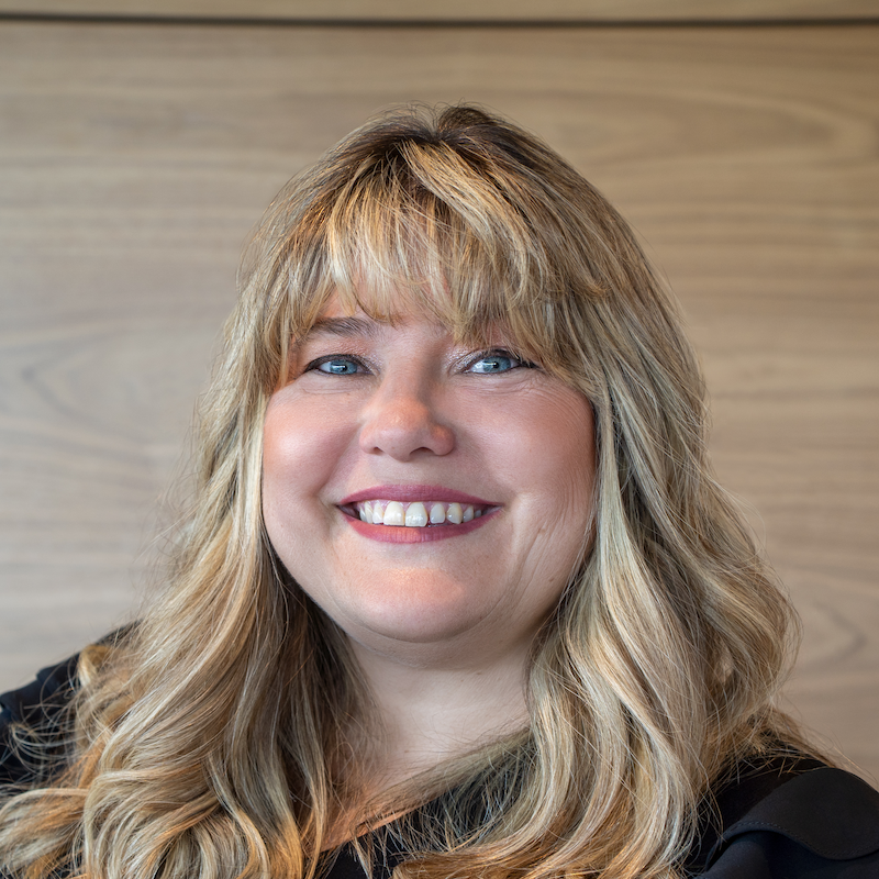 A smiling woman with long wavy blonde hair, blue eyes, and wearing a black top, standing against a wooden background.