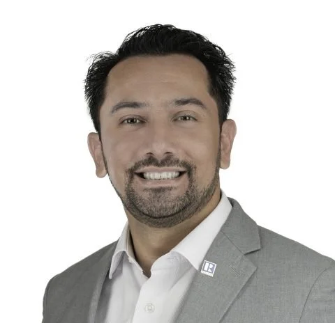 Professional headshot of a man with dark hair, beard, smiling, wearing a light gray suit and white shirt against a white background.