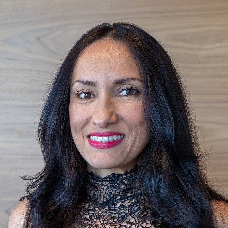 Close-up of a woman with long dark hair, smiling, wearing a black lace top, standing against a light wood background.
