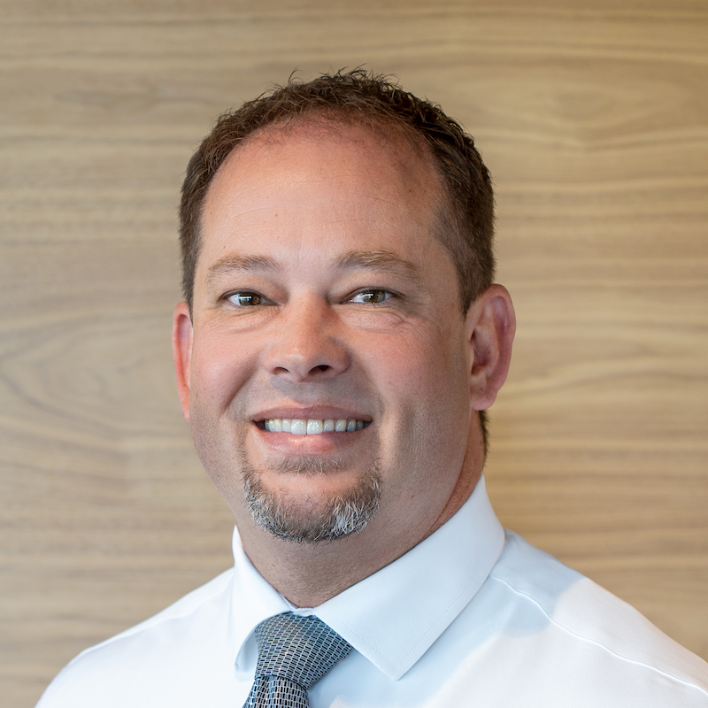 Close-up portrait of a smiling man with short brown hair, a goatee, wearing a white dress shirt and a gray tie, standing in front of a wooden background.