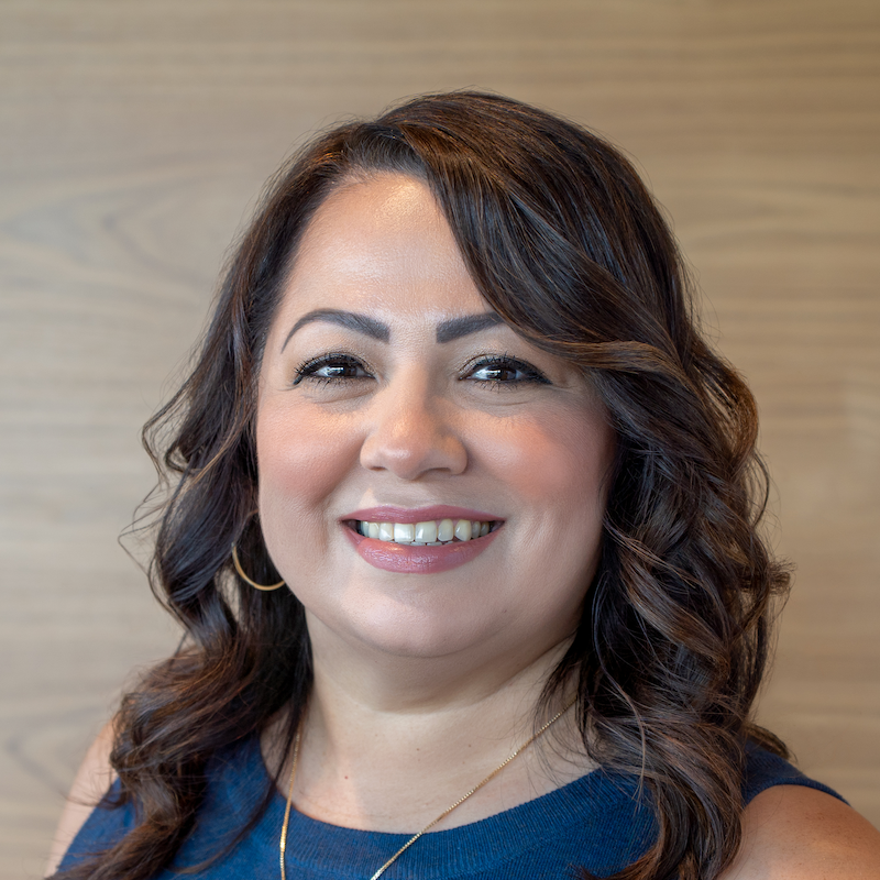 A woman with shoulder-length dark brown, wavy hair, wearing a navy sleeveless top, gold hoop earrings, and a delicate gold necklace, smiling in front of a wooden background.