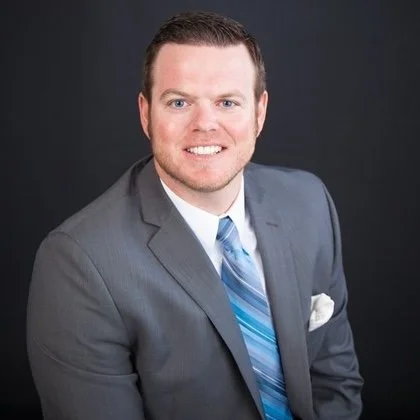 Professional man in a gray suit and blue striped tie smiling against a dark background.