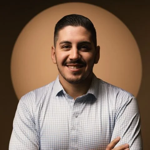 Portrait of a smiling man with short dark hair and a trimmed beard, wearing a light checkered button-up shirt, standing in front of a neutral background.