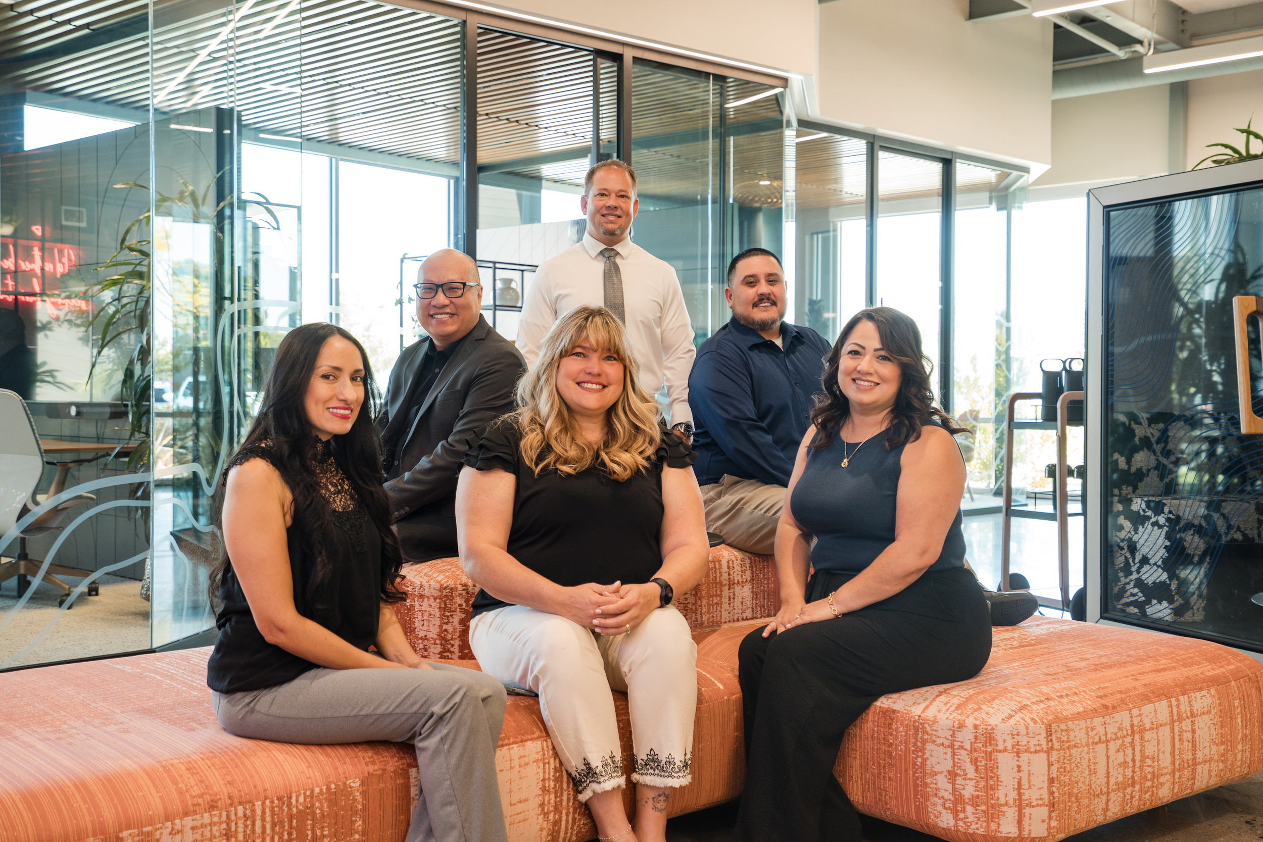 A group of seven diverse professionals, five women and two men, posing together in a modern office lobby with glass walls and indoor plants.