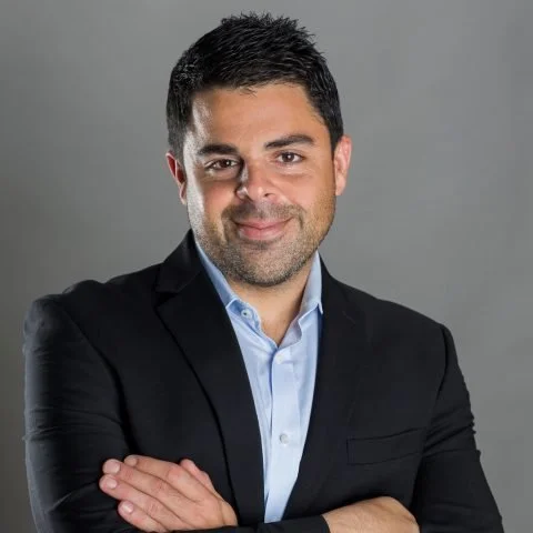 Portrait of a man with dark hair, wearing a black suit jacket and a light blue shirt, smiling with arms crossed against a gray background.